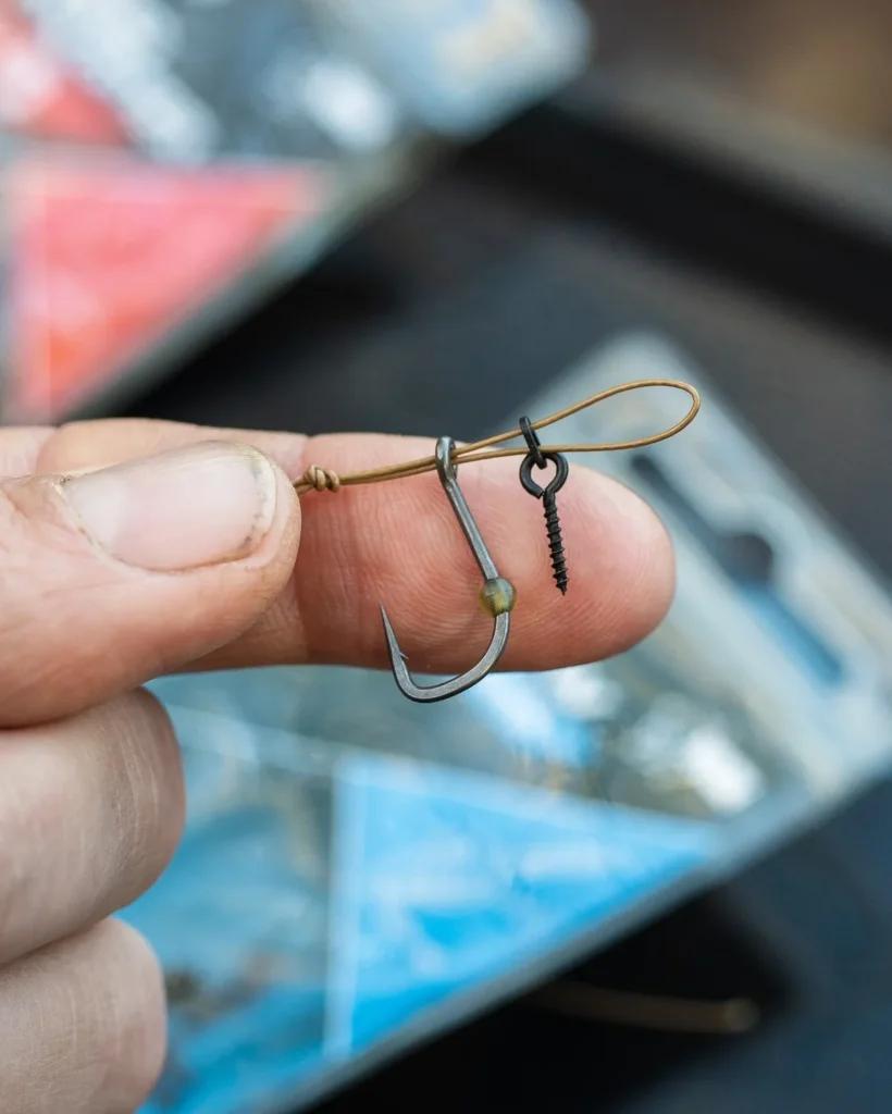 Carp hook with a multi rig setup showing the D-loop and bait screw attachment, held between fingers with tackle packets blurred behind.