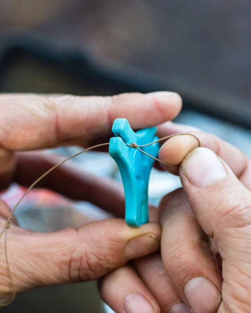 Hands holding a small rig tool while tightening a knot in coated hooklink material for a carp rig, with tackle tray in the background.