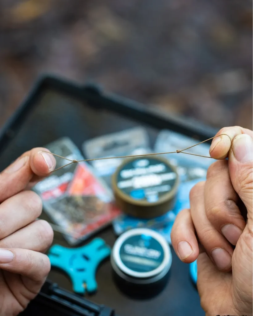 Close-up of a carp hook and multi rig attachment being assembled, with the D-loop and bait screw visible, held between fingertips.
